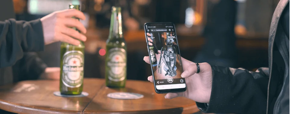 Celular mostrando a taça da Champions League em realidade aumentada, ao lado de uma Heineken